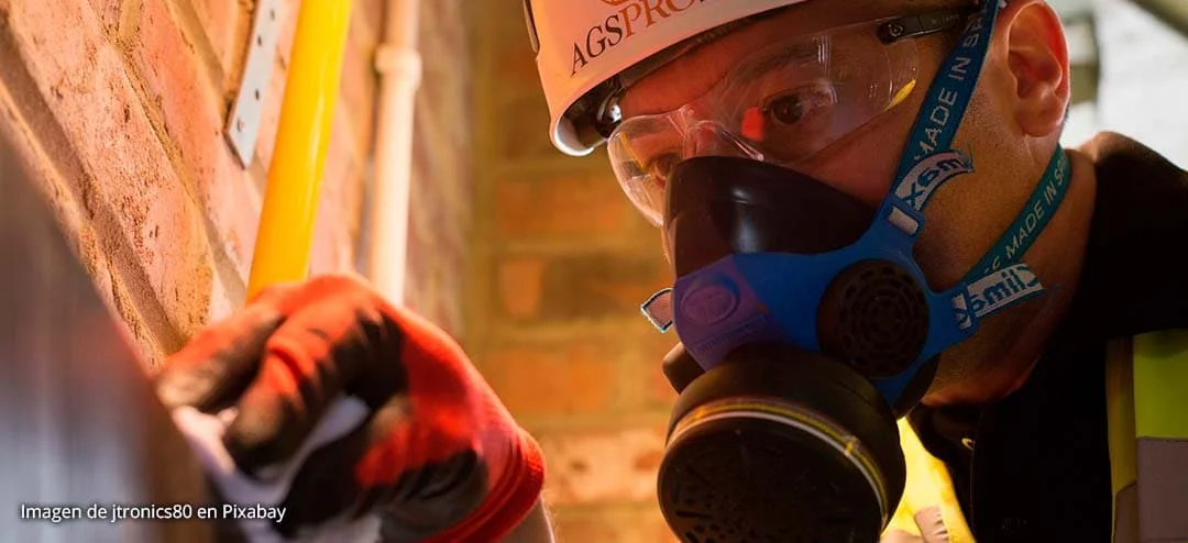 Portada publicaciones-pi2 Trabajador con mascarilla y casco industrial inspecciona superficie metálica en entorno cerrado